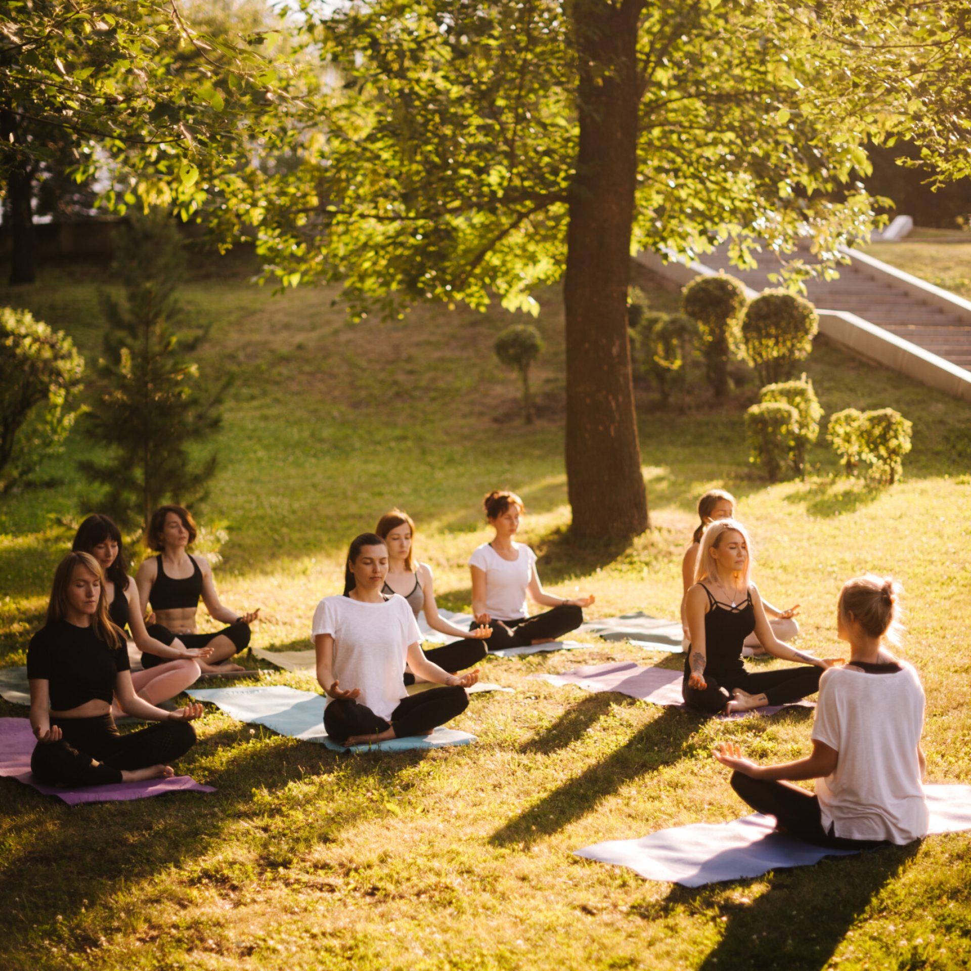 Group of young women meditate in city park on summer sunny morning under guidance of instructor. Group of peaceful people are sits and meditate outdoors in lotus pose on grass with closed eyes