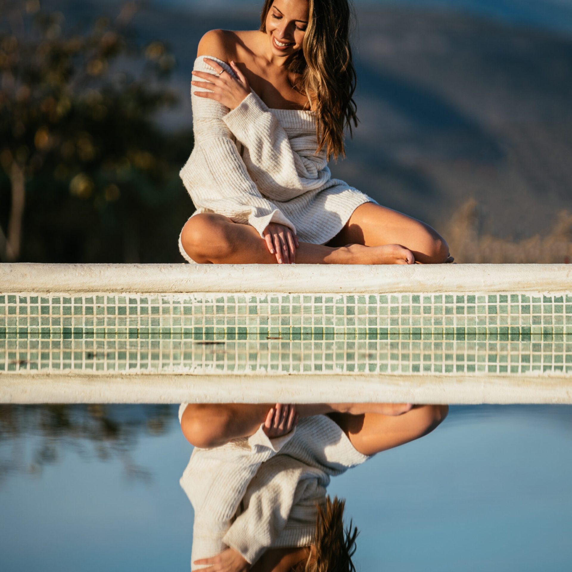 Positive female in soft sweater smiling and closing eyes while relaxing on border in yard on blurred background of mountains